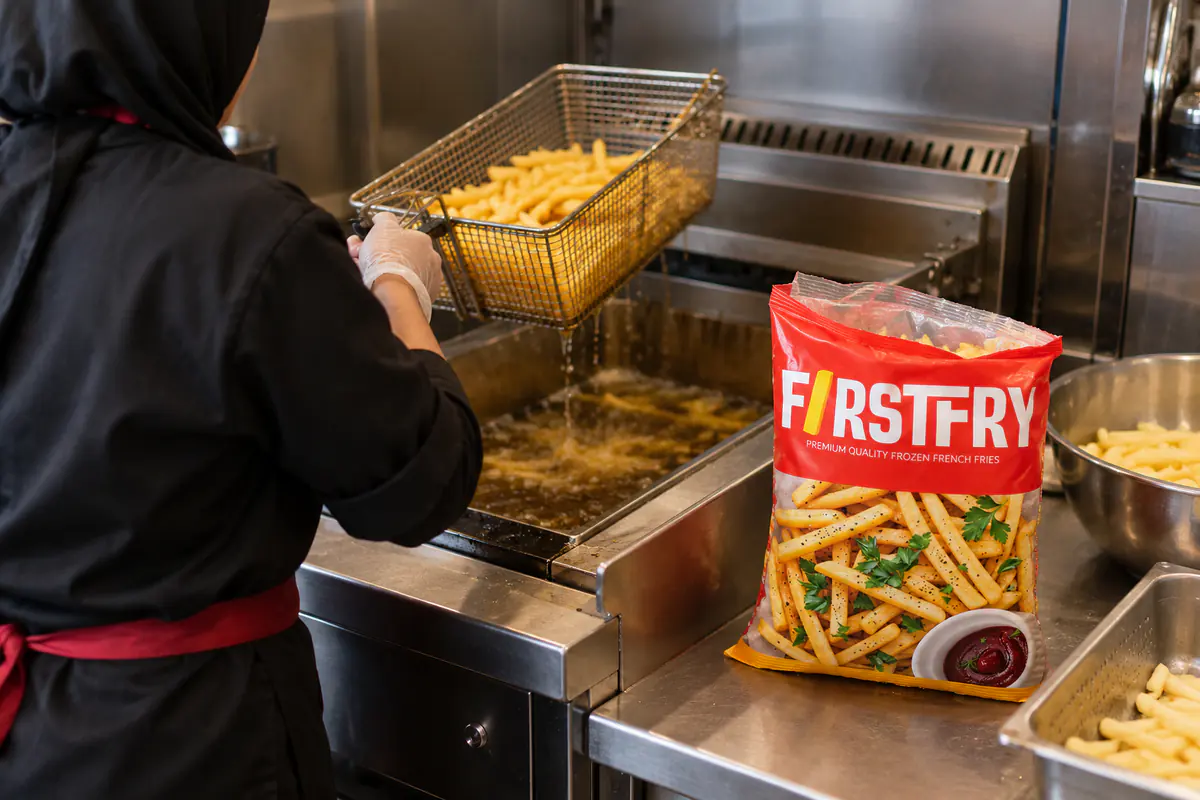 A kitchen worker in a headscarf lifting a fry basket beside an open FirstFry pack
