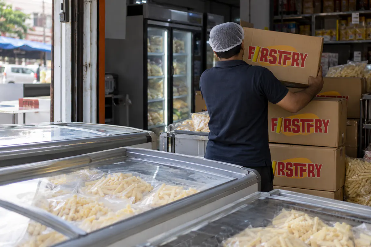 A shopkeeper seen from behind lifting a FirstFry carton at an open-fronted shop